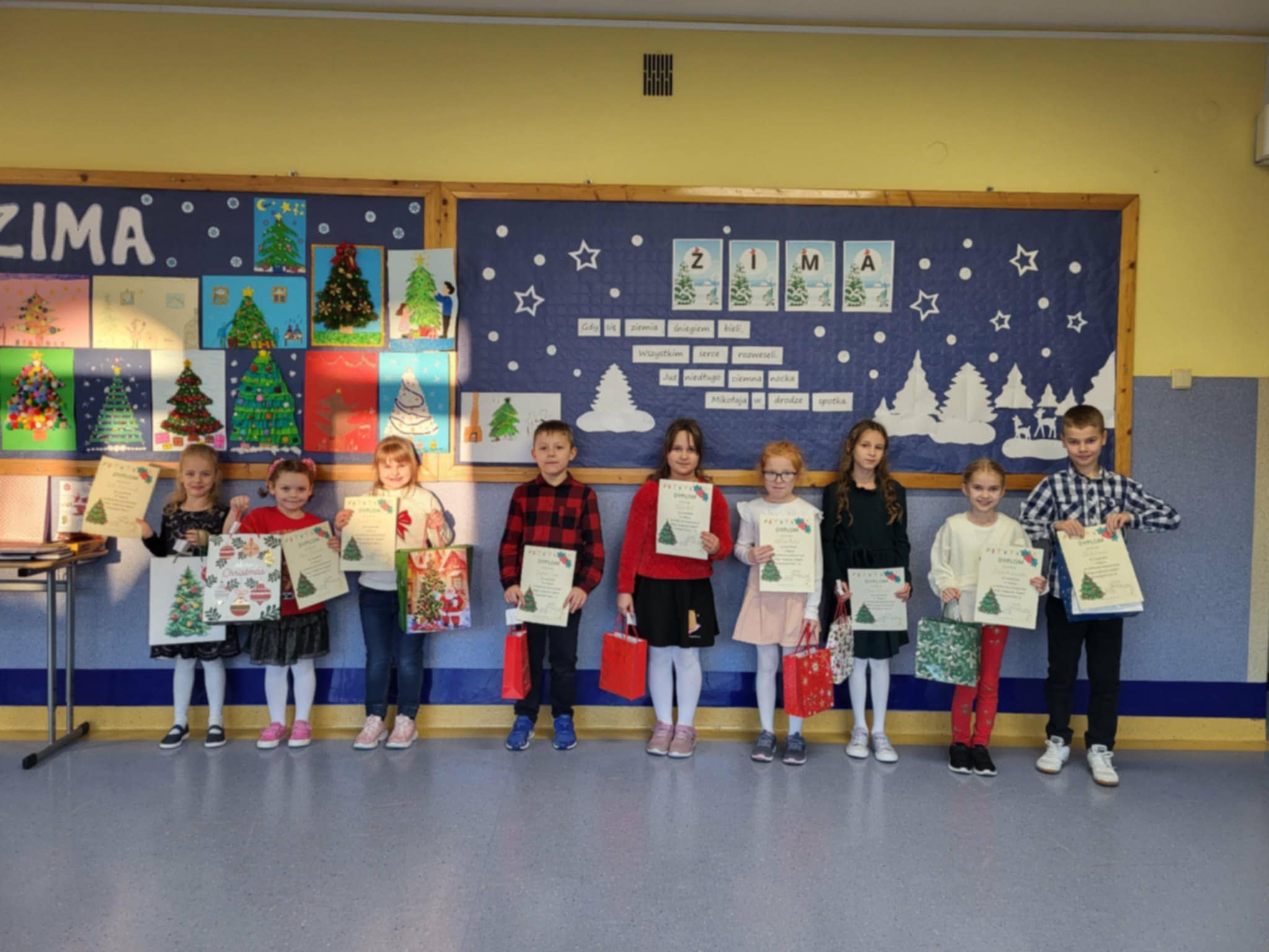 Uczniowie klas 0–3 stoją w rzędzie na szkolnym korytarzu, trzymając dyplomy i drobne upominki. Za nimi widoczna jest gazetka ścienna o tematyce zimowo-świątecznej z pracami plastycznymi przedstawiającymi choinki.