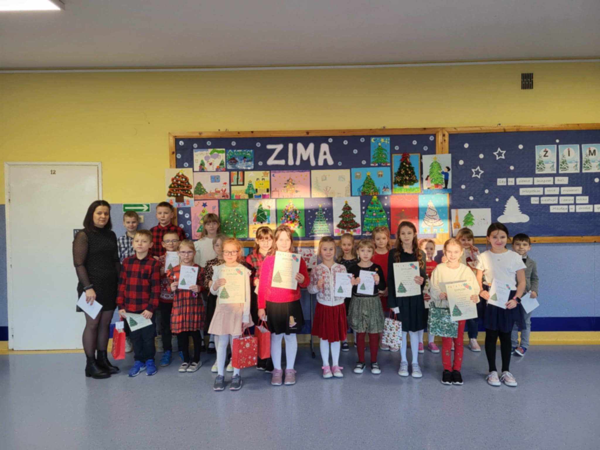 Grupa uczniów wraz z nauczycielem pozuje do wspólnego zdjęcia na szkolnym korytarzu. Dzieci trzymają dyplomy i upominki, a w tle znajduje się wystawa prac plastycznych o tematyce zimowej i świątecznej.