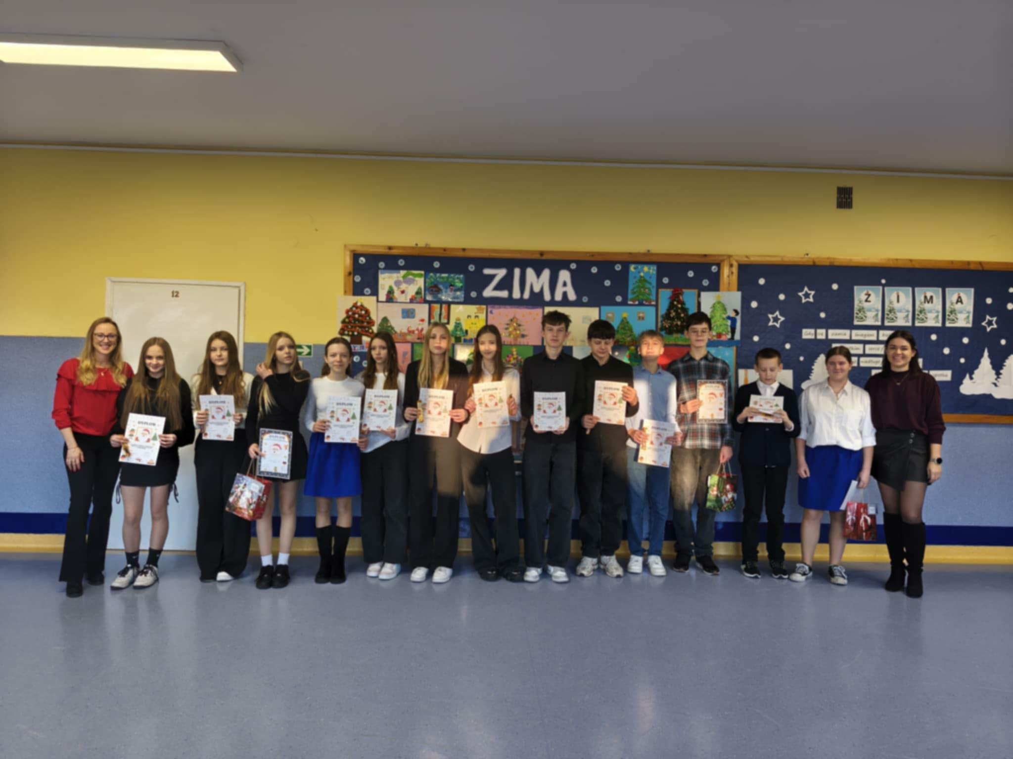 Zdjęcie grupowe uczniów wraz z nauczycielami, pozują do wspólnego zdjęcia na szkolnym korytarzu. Dzieci trzymają dyplomy i upominki. 