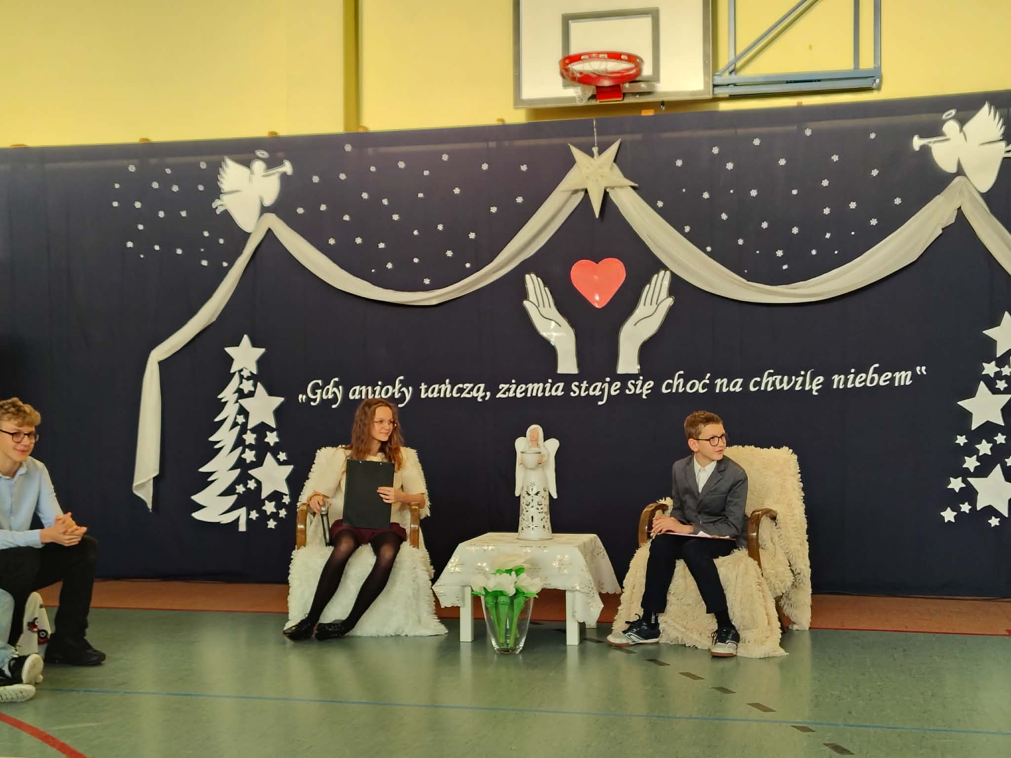 Uczniowie prowadzący apel siedzą na fotelach, w tle dekoracja świąteczna.