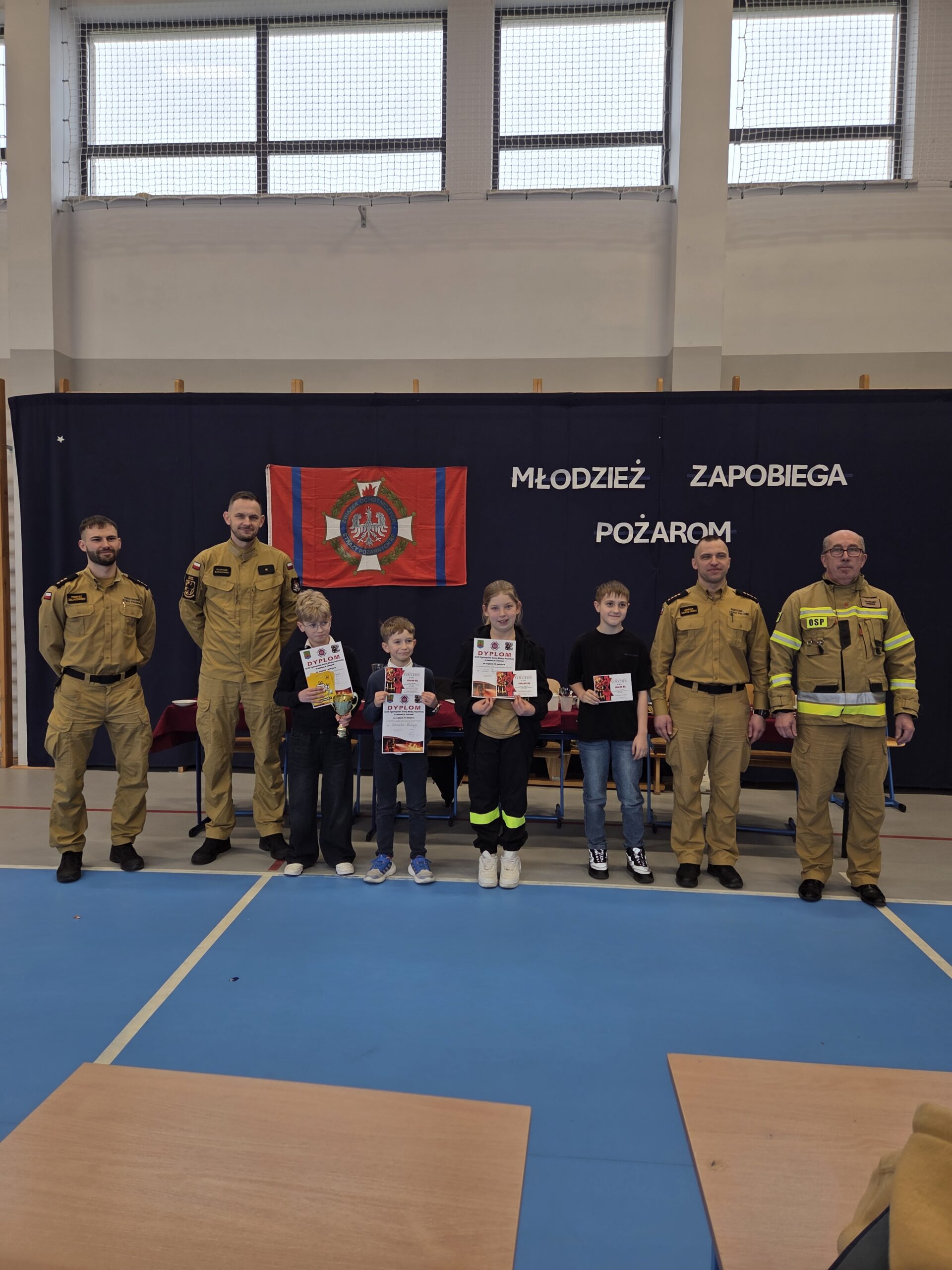 Zdjęcie grupowe uczniów i komisji podczas rozdania nagród. W tle gazetka Młodzież Zapobiega Pożarom.
