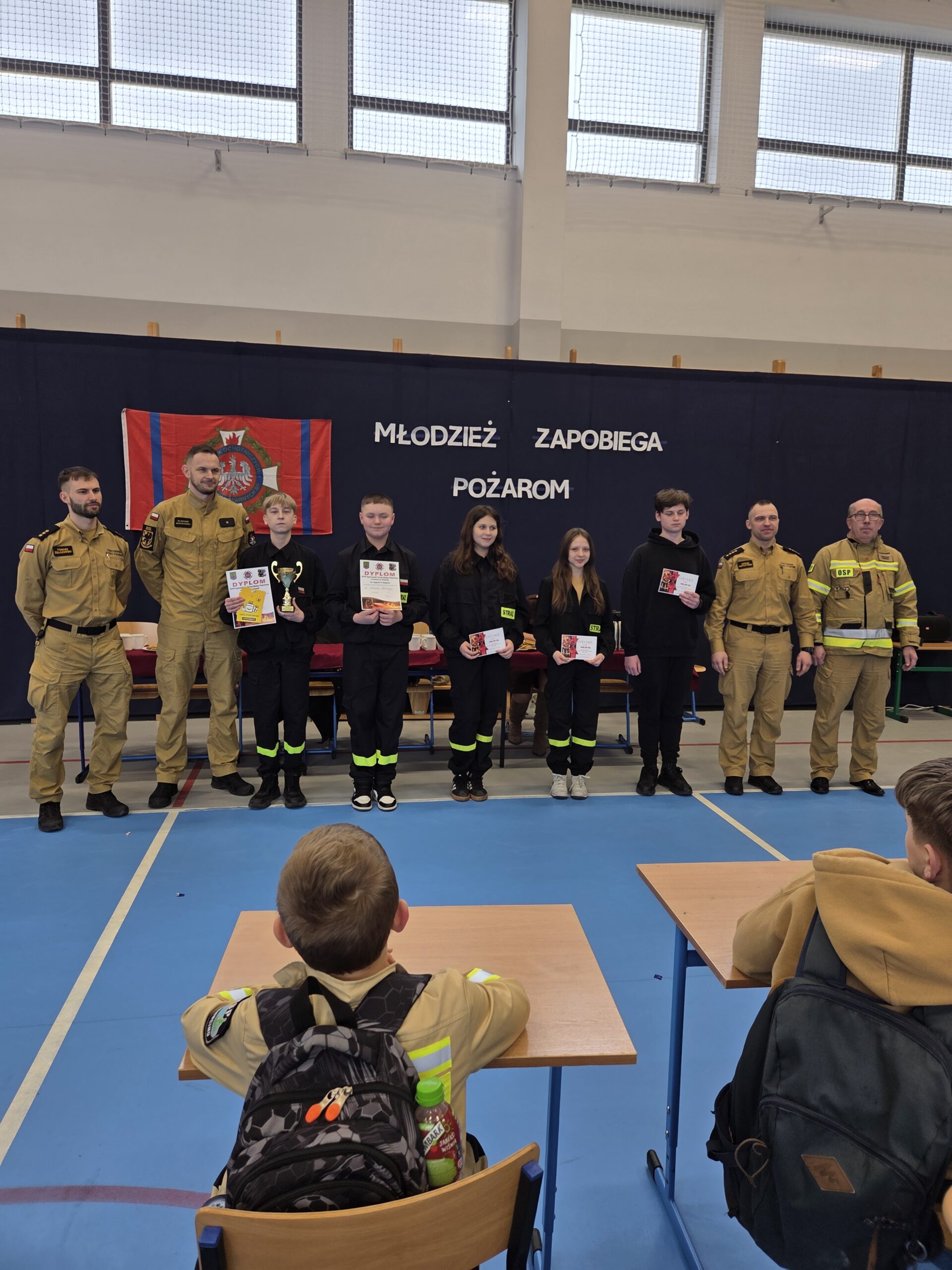 Zdjęcie grupowe uczniów ze starszej kategorii i komisji podczas rozdania nagród. W tle gazetka Młodzież Zapobiega Pożarom.