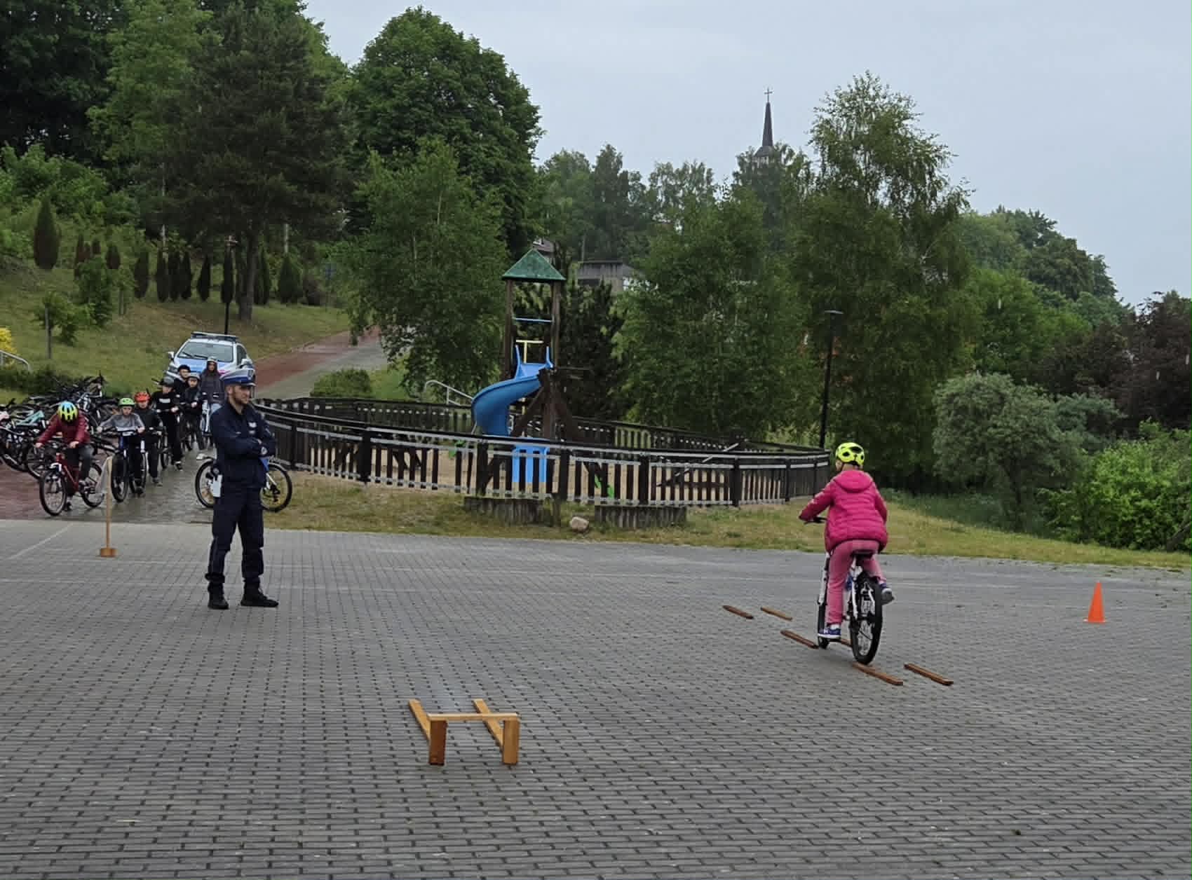 Uczennica na rowerze pokonuje plac manewrowy a Pan Policjant sprawdza poprawność wykonania.