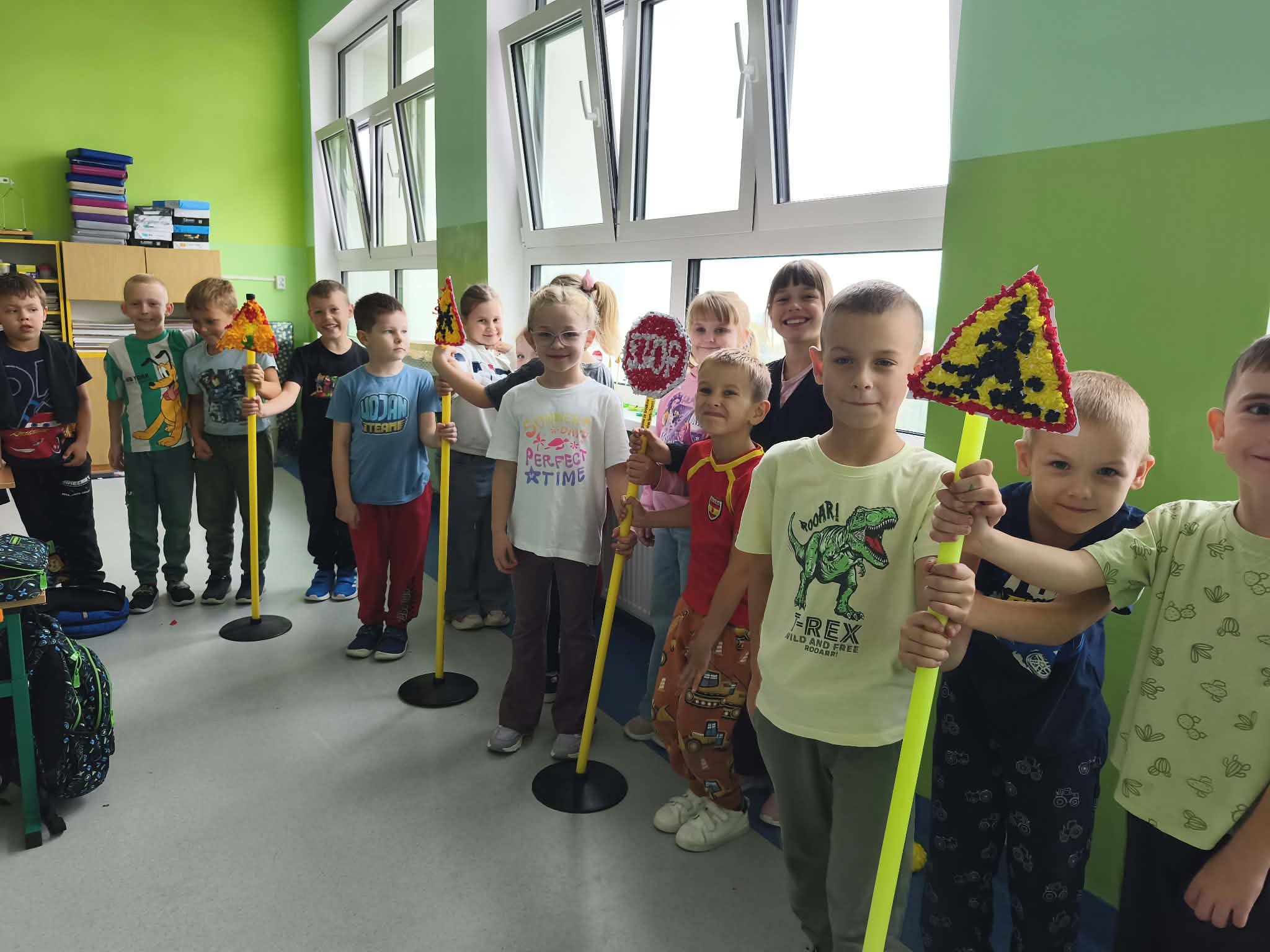 Grupa dzieci w sali szkolnej prezentuje wykonane przez siebie makiety znaków drogowych. Dzieci trzymają kolorowe znaki na długich tyczkach.