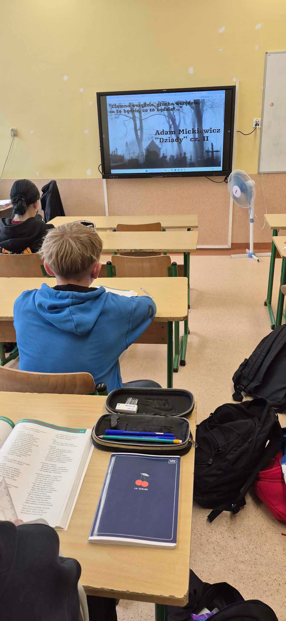 Uczniowie w sali lekcyjnej siedzą w ławce i czytają Dziady.