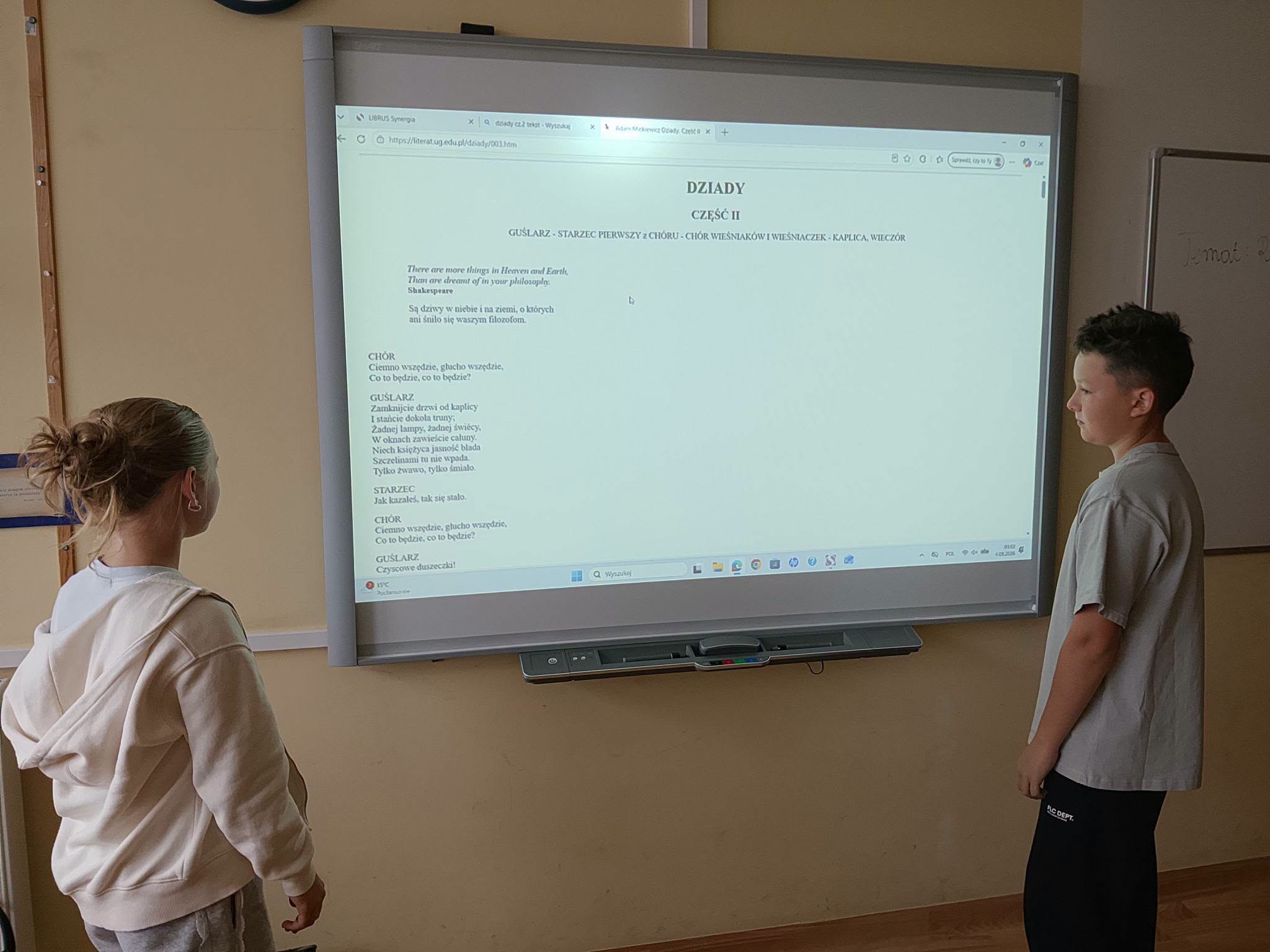 Dwóch uczniów stoi przed tablicą interaktywną i czyta na głos teks z II części Dziadów.